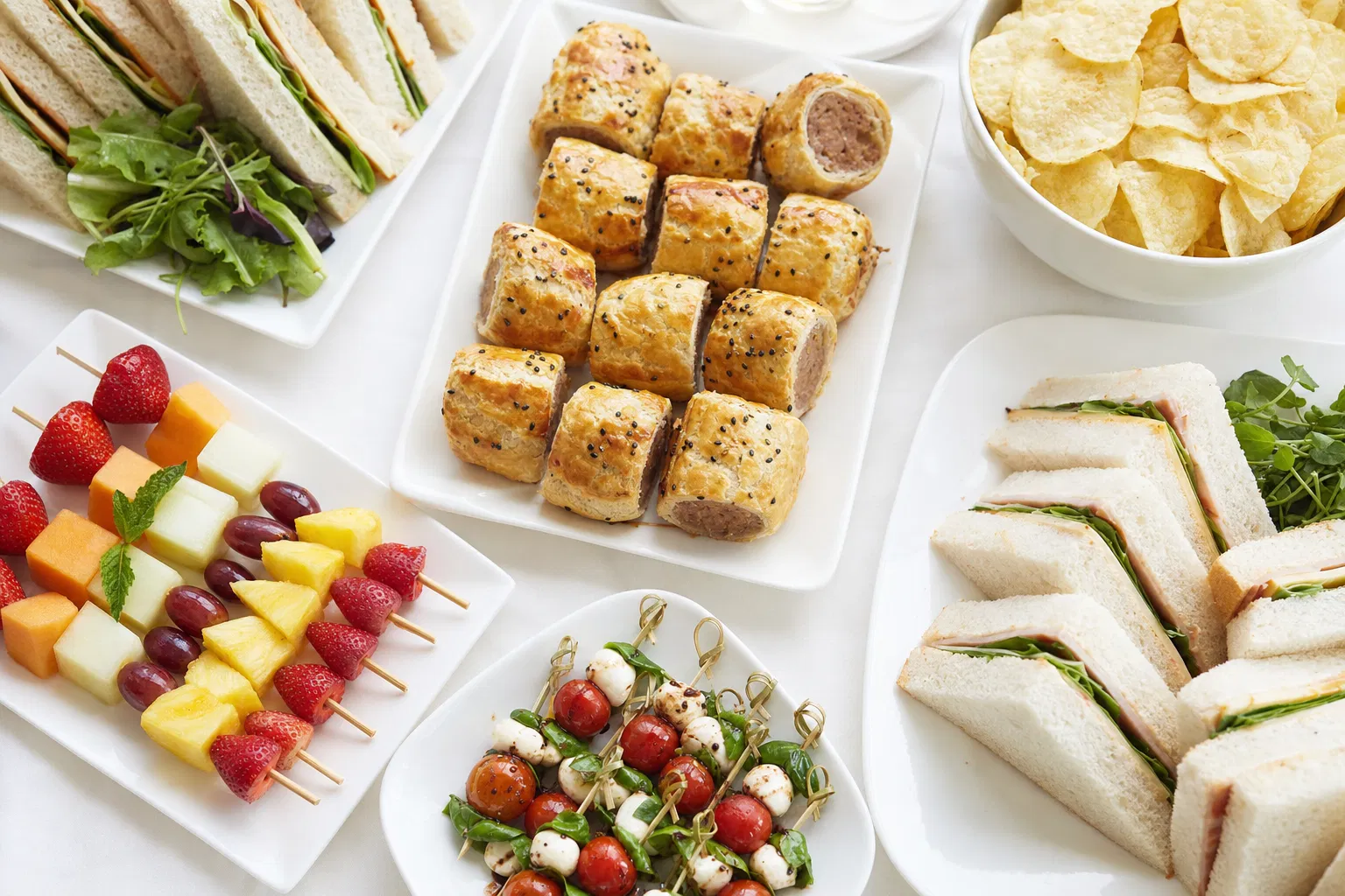 Catering buffet spread with sandwiches, sausage rolls, and fruit kebabs